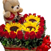 Canasta cuadrada con rosas rojas dispuestas en forma de corazón y girasoles en centro con tierno oso de peluche.